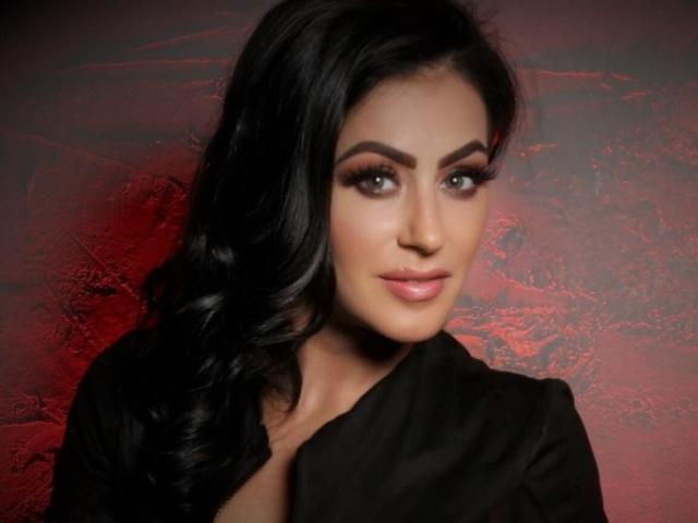 Photograph of MistressTaylor, dark-haired woman with sultry eyes, full lips, wearing a black top. Red textured background. Sexual tension, looking directly at camera, slight smirk. Slight glow on face.