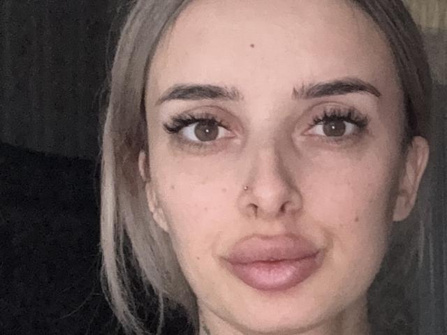 Close-up of KatrinaHotX69 with pale skin, brown eyes, and full lips. She has light brown hair, a nose piercing, and long eyelashes. Background is dark. Fucking sexy, looking straight at camera.