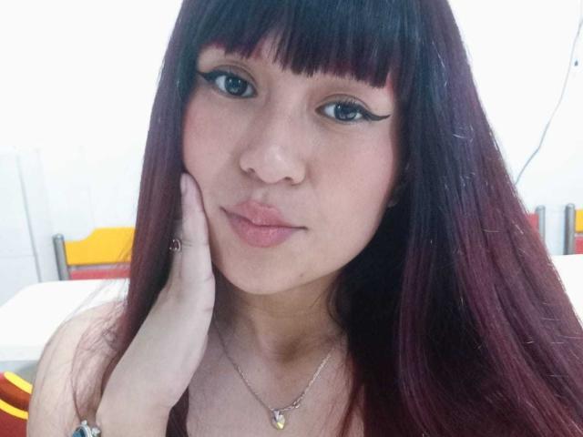 Nahaufnahme von JadeMadiysson, einer asiatischen Frau mit langen, dunkelbraunen Haaren und Pony, die einen auffälligen, schwarzen Eyeliner trägt. Sie legt ihre Hand auf ihre Wange und schaut direkt in die Kamera. Weiches Licht betont ihr Gesicht..