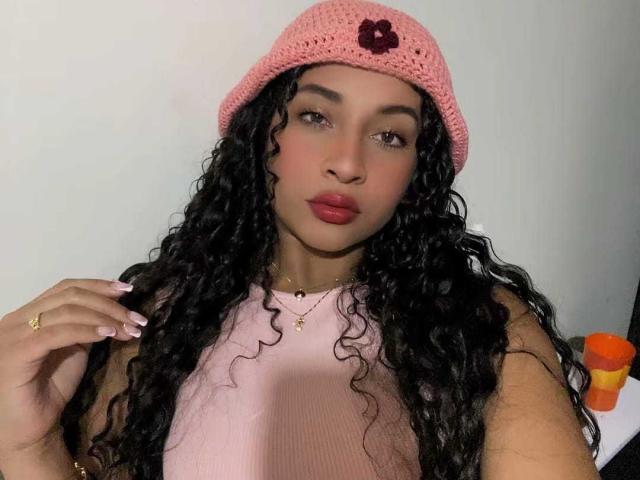 Fotografia de HollySwett, com pele morena e cabelos longos e cacheados pretos, usando um gorro rosa de tricô e uma blusa rosa canelada, em corpo inteiro. lábios vermelhos, segurando o cabelo com um anel de ouro, olhando para a câmera.