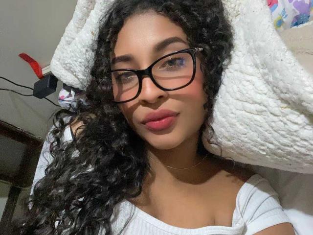 Holly Swett, eine attraktive lateinamerikanische Frau mit langen, lockigen schwarzen Haaren, trägt eine schwarze Brille und einen weißen Hut und blickt verführerisch. die Kamera mit rosa Lippenstift, in einem weißen Oberteil.