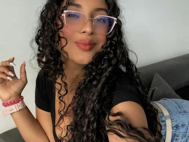 Fotografía de HollySwett con cabello negro largo y rizado, usando gafas de color rosa, una blusa negra y jeans azules. Ella sostiene un cigarrillo, se lame los labios con una mirada seductora, y lleva un reloj rosa..