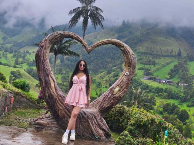 Holly Swett está de pie frente a una gran escultura de madera con forma de corazón, vestida con un vestido rosa y botas blancas, con el pelo largo. cabello negro. Colinas verdes exuberantes y un cielo brumoso en el fondo..