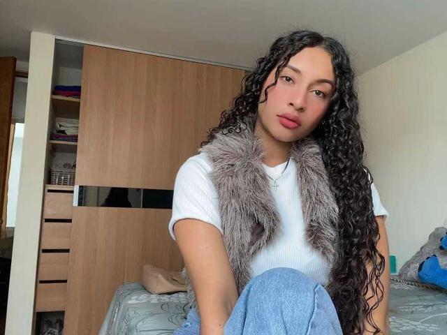 Holly Swett, con cabello negro largo y rizado, vestida con una camiseta blanca y una falda azul, lleva un chaleco gris y peludo. Ella está sentada en una cama, y en el fondo se ve un armario de madera. Increíblemente atractivo/a..