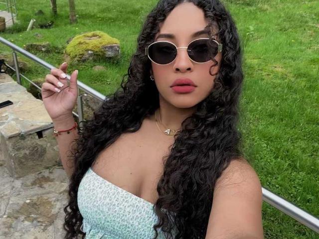 Fotografía de HollySwett, con cabello negro largo y rizado, usando gafas de sol redondas, un top blanco sin tirantes y un collar. Ella sostiene un cigarrillo, está de pie al aire libre, cerca de una zona con césped, y tiene una expresión seductora..