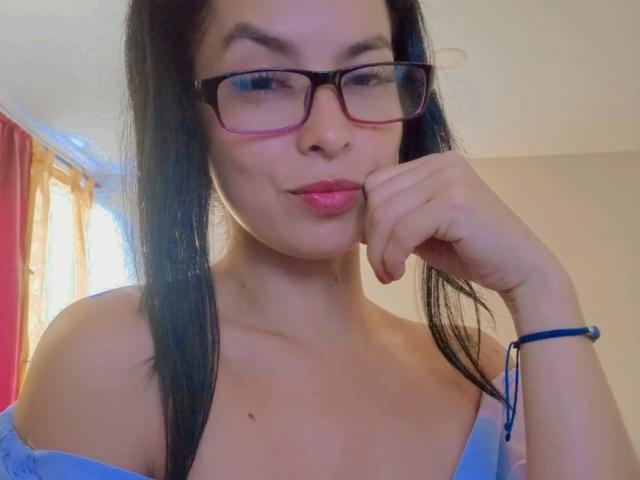 Primo piano di Kathelott, una donna con lunghi capelli neri, che indossa occhiali con montatura rosa e una maglietta blu a spalle scoperte. appoggiando il mento sulla mano, mostrando un'espressione sexy e sicura di sé..
