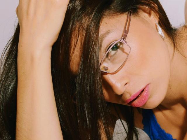 Fotografia ravvicinata di Kathelott, una donna attraente con lunghi capelli neri, che indossa occhiali e una maglietta blu, con una mano tra i capelli. occhi verdi, rossetto rosa, sguardo intenso, testa leggermente inclinata.