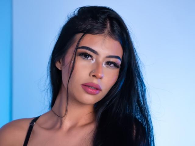 Foto van AmilyBella, een aantrekkelijke vrouw met lang zwart haar, doordringende bruine ogen, roze lippenstift en een neusring. Ze draagt een zwarte top met bandjes, op een lichtblauwe achtergrond. Ongelooflijk mooi..