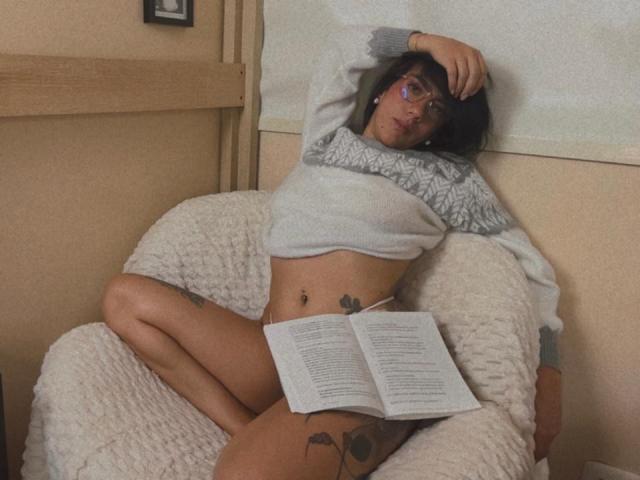 Dominatrix Melissa, desnuda excepto por un suéter gris, se recuesta en un sillón blanco con textura, con las piernas separadas, leyendo un libro, gafas, tatuajes en muslos y abdomen, brazo levantado, relajada, sexy.