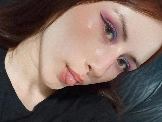 Close-up of MalenaJones, a fair-skinned woman with dark brown hair, purple eyeshadow, and a nose piercing. She's looking at the camera with a slight, pouty expression. Fucking sexy.