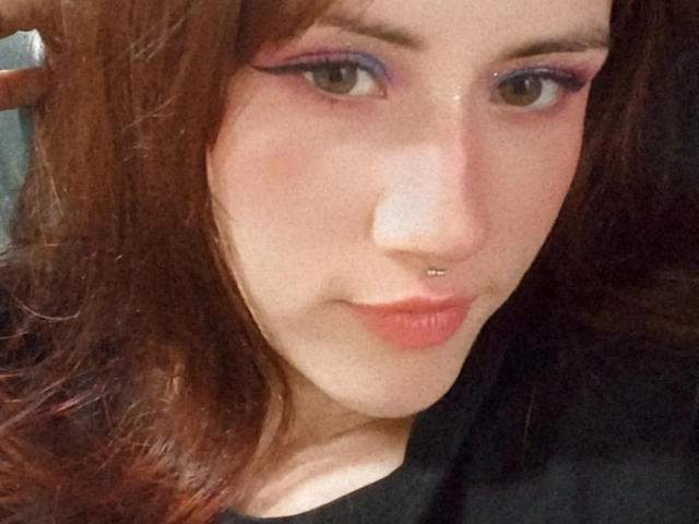 Close-up of MalenaJones, fair-skinned with red-brown hair, purple eyeshadow, and a nose piercing. She has pouty lips, wearing a black top. Sexual tension, seductive gaze, slight smirk.