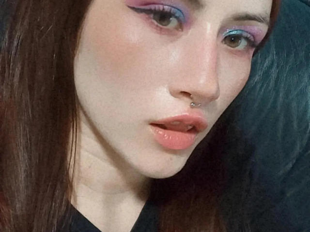 Close-up of MalenaJones, a pale-skinned woman with dark brown hair, purple eyeshadow, and a silver nose ring. She has parted lips, slightly parted, looking directly at the camera. Her expression is seductive.