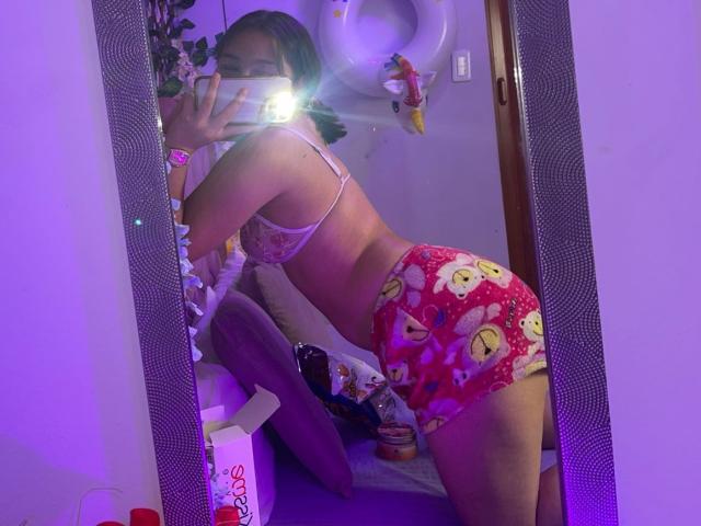 Emma Quintana est agenouillée devant un miroir, en train de prendre un selfie avec le flash. Elle porte un soutien-gorge en dentelle et un short de pyjama à fleurs rouges, mettant en valeur ses jolies formes. Éclairage violet. Reflet dans un miroir.