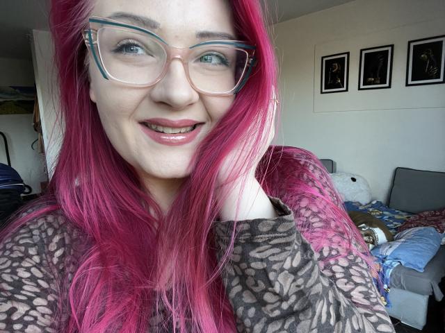 Close-up of RedKissRuby with red hair, wearing clear glasses and a black lace top. She's smiling, with sunlight highlighting her face, and three framed pictures on the wall behind her.