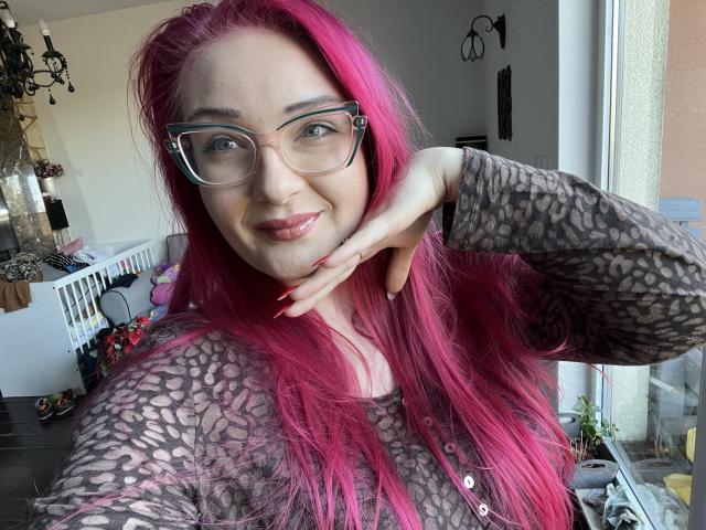 Photographie de RedKissRuby avec des cheveux roses éclatants, portant des lunettes et un haut en dentelle noire, souriant de manière séduisante. Une main sur sa joue, une lumière vive venant d'une fenêtre, une ambiance séduisante..