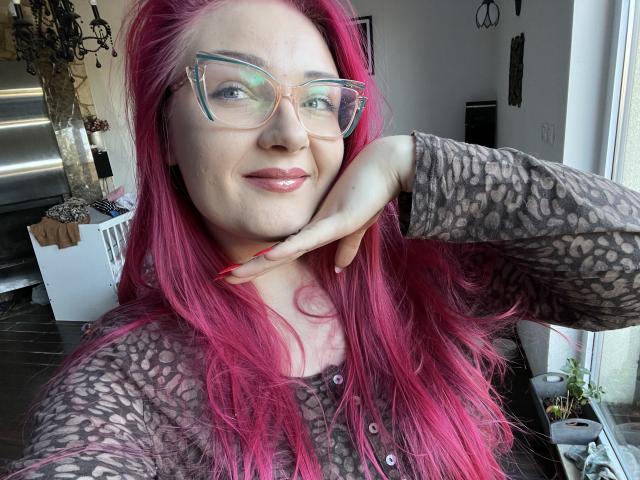 Photograph of RedKissRuby with long, bright pink hair, wearing clear glasses and a black lace top, touching her chin with a hand. She's smiling subtly, exuding confident sexy vibes.