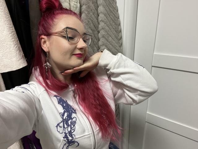 RedKissRuby, met rood haar in een hoge paardenstaart, draagt een bril en een witte hoodie met blauwe designs, en poseert verleidelijk. Hand op de nek, witte kast en grijze handdoek op de achtergrond..