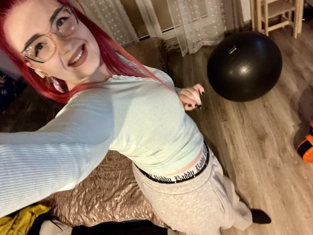RedKissRuby, una donna formosa con capelli rossi e occhiali, si scatta un selfie dall'alto. Indossa una maglietta bianca e una gonna beige, sta sorridendo, e sullo sfondo si vede una palla da ginnastica nera..