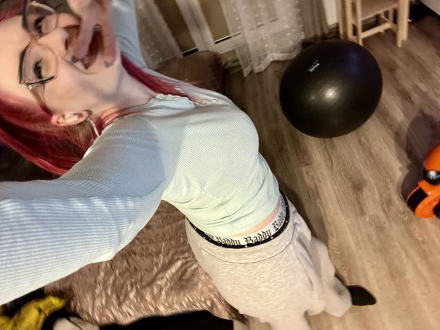 Nahaufnahme eines Fotos von RedKissRuby, einer rothaarigen Frau mit Brille, in einem weißen Rippstrickoberteil, einem weißen Rock und einem schwarzen Gürtel, die sich zurücklehnt. Augen halb geschlossen, schwarzer Trainingsball und ein orangefarbenes Objekt auf Holzboden..