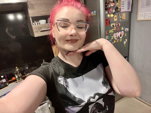 RedKissRuby met roze haar en een bril, een zwart-wit T-shirt, hand op kin, licht glimlachend, keuken op de achtergrond. Koelkast beplakt met stickers, zachte verlichting, casual selfie. Heel schattig.