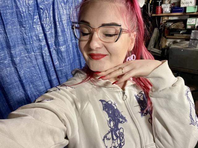 RedKissRuby, une femme à la peau claire avec des cheveux roux, portant des lunettes et un sweat-shirt blanc avec un motif bleu, qui la touche. cou, sourire narquois, rideau bleu en arrière-plan, pose légèrement suggestive.