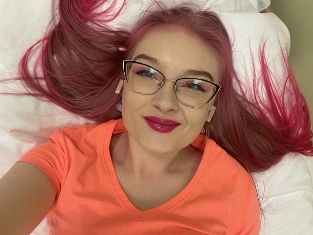 RedKissRuby ligt op een wit bed, gekleed in een feloranje T-shirt en een bril, met roze haar dat uitgespreid is. Rode lippenstift, een lichte glimlach, creëert een sexy en ontspannen sfeer..
