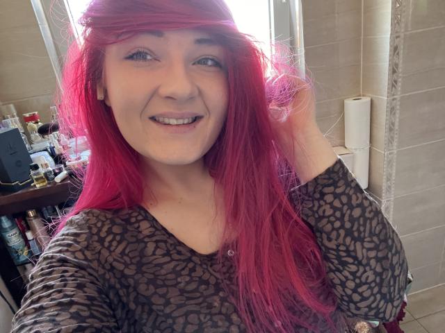 RedKissRuby, una donna con i capelli rosso acceso, sorride alla macchina fotografica. Indossa una maglietta nera con un motivo, ed è in piedi in un bagno. La luce del sole illumina i suoi capelli, creando un aspetto seducente e invitante..