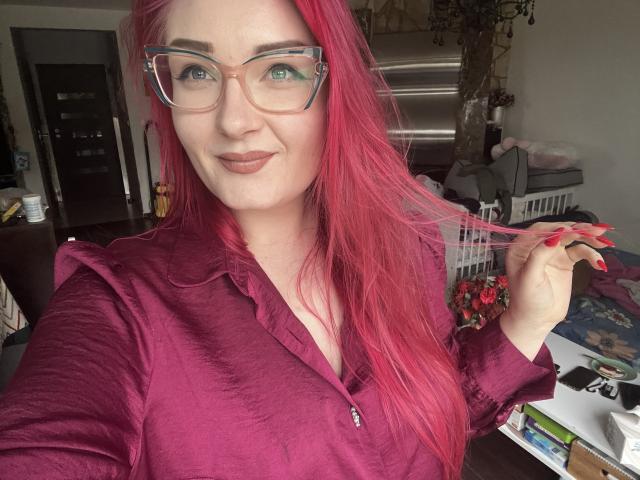 RedKissRuby, con i suoi lunghi capelli rossi e gli occhiali chiari, tiene una ciocca di capelli con la mano destra. Indossa una camicetta color borgogna, sorridendo leggermente, con una scrivania disordinata sullo sfondo..