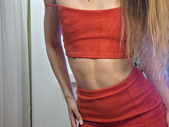 Photograph of a slim, sexy woman with long blonde hair, wearing a tight red crop top and matching high-waisted skirt. Her hand grips her hip, emphasizing her flat stomach and curves.