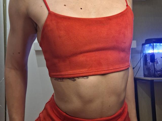 Fotografía de primer plano de MilaYanis, que lleva un top corto rojo, revelando un abdomen tonificado y delgado. Pantalones cortos rojos, acuario azul con luces brillantes en el fondo. Tensión sexual, pose casual..