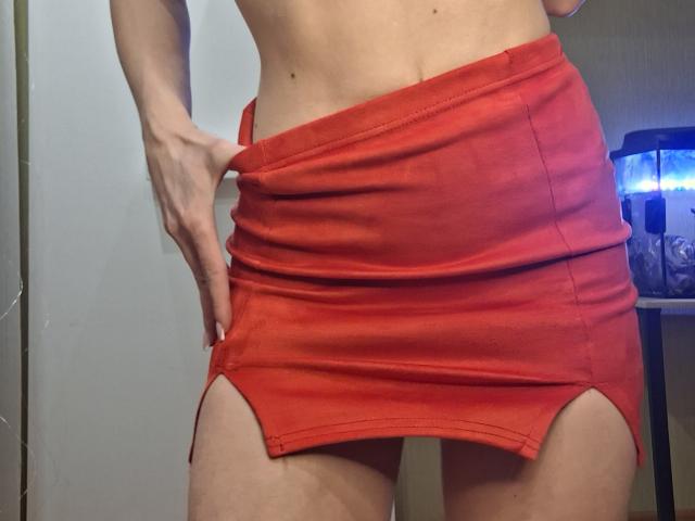 Fotografía de primer plano de la parte inferior del cuerpo de MilaYanis, vistiendo una minifalda roja ajustada con aberturas laterales, con una mano tirando de la falda hacia abajo. revela su cadera. Luz brillante proveniente del lado derecho.