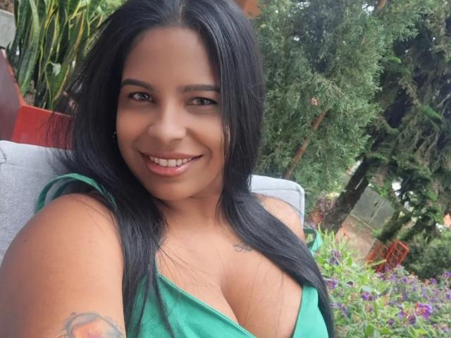 Fotografia de Chloe Harrison com cabelos longos e pretos, sorrindo para a câmera. Ela está vestindo uma blusa verde com decote baixo, que revela seios grandes. Cenário ao ar livre com árvores e flores coloridas ao fundo..