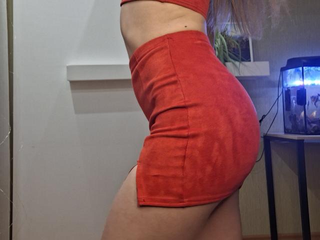 MilaYanis vestía una minifalda roja ajustada y una blusa a juego, vista de perfil, que resaltaba sus curvas, sus glúteos tonificados y su cintura delgada. La tela roja se adhiere a su piel, resaltando su figura sexy y curvilínea. Acuario en el fondo.