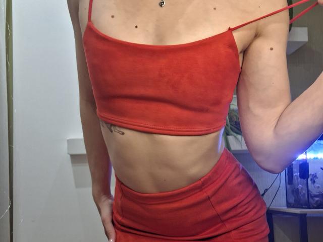 Photographie d'une femme mince, à la peau claire, avec de petits seins, portant un débardeur rouge moulant et un pantalon taille haute assorti. jupe. Sa main droite repose sur sa hanche, révélant un tatouage. Elle porte un petit pendentif autour du cou. Son haut rouge est légèrement baissé, dévoilant une partie de son ventre. Sa jupe est légèrement froissée, ce qui met en valeur ses courbes..