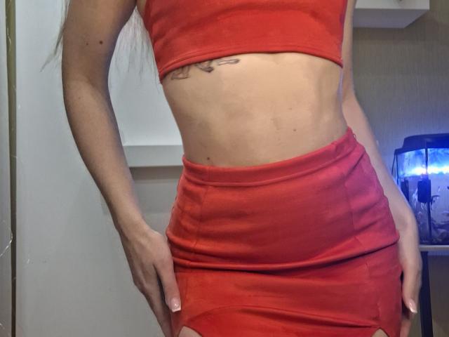 Fotografía de una mujer, MilaYanis, que lleva un top corto rojo ajustado y una falda roja a juego, de pie con las manos en las caderas. resaltando su cintura delgada y tonificada, así como su tatuaje visible. Iluminación brillante.