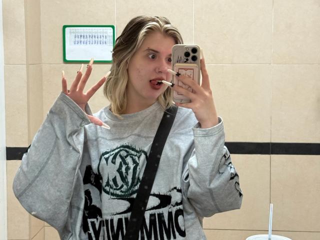 Lennie Caronna, una giovane donna con i capelli biondi, si scatta un selfie allo specchio in un bagno molto luminoso. Indossa una felpa grigia con la scritta "Evil Mind" e alza la mano destra. iPhone in mano. Tensione sessuale.