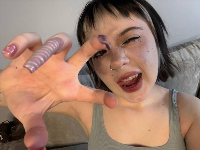 Photo en gros plan de LennieCaronna, cheveux noirs courts, peau claire, taches de rousseur, la langue tirée, tenant un godemiché violet texturé. main droite, portant un débardeur gris, doigts écartés, expression sexuelle intense..