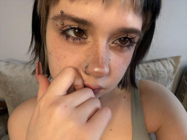 Close-up de Lennie Caronna, uma jovem com cabelo preto curto, sardas e maquiagem escura nos olhos. Ela está mordendo o dedo, olhando de forma sedutora com olhos brilhantes e cintilantes, vestindo uma camiseta cinza..