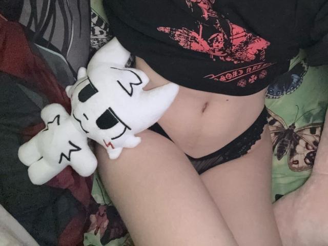 Close-up photo of LennieCaronna's lower body, wearing black lace panties and a black t-shirt with a red graphic. A white plush toy rests on their hip. Pale skin, legs slightly spread.