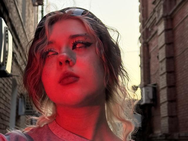 Foto em close-up de LennieCaronna, com a cabeça ligeiramente inclinada, iluminação vermelha que projeta sombras intensas no rosto, cabelo loiro ondulado. maquiagem pesada nos olhos, fundo de um beco urbano, edifícios de tijolos, forte tensão sexual, olhar sedutor..