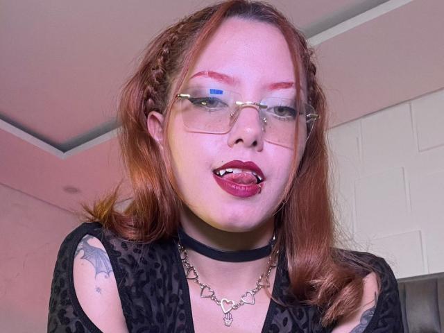 Close-up of EmmaThomsonn with red hair, braided crown, wearing glasses, black choker, black lace top, tongue out, red lipstick, tattoo on left shoulder, white walls in background. Fucking sexy.