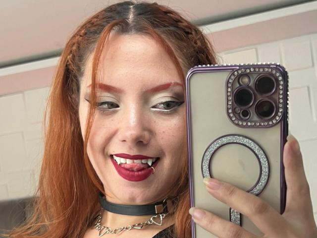 Close-up foto van EmmaThomsonn met rood haar, sproeten en donkere lipstick, die haar tong uitsteekt terwijl ze een telefoon vasthoudt. met een behuizing met een rand. Ze draagt een zwarte choker en een ketting..