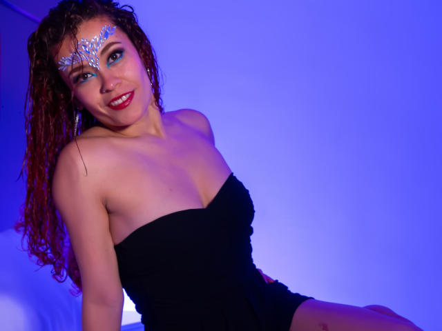 Arielle Brooks mit nassen, lockigen braunen Haaren, die ein schwarzes, trägerloses Kleid und eine silberne Gesichtsmaske trägt, lächelt und hat rote Lippen. Lippenstift. Blauer Hintergrund, nach vorne gebeugt, Ausschnitt betont, anzügliche Pose..
