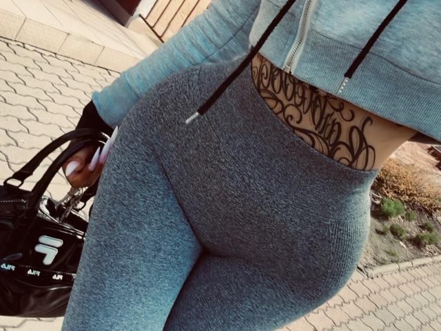 Primo piano delle curve del sedere di LuxuryDoll69, in leggings grigi aderenti, che mostra un tatuaggio nero sulla parte bassa della schiena. Indossa una felpa grigia chiara e porta una borsa nera nella mano destra..