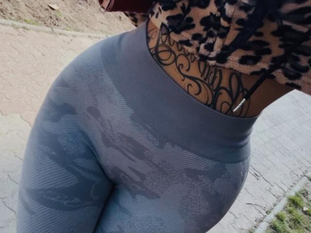 Primo piano della parte inferiore del corpo di LuxuryDoll69, con particolare attenzione ai suoi leggings grigi mimetici, aderenti e che valorizzano le sue curve. grande e rotonda. La sua maglietta a stampa leopardo rivela un intricato tatuaggio nero sulla vita..