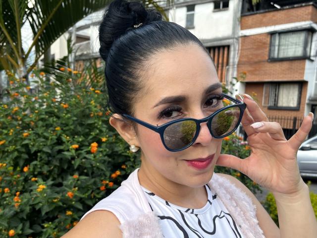 Fotografia di Katty Rivera, una donna attraente con i capelli scuri raccolti in uno chignon, che indossa occhiali da sole blu, una camicetta bianca in pizzo e rossetto rosso.
lei sta toccando gli occhiali da sole con un sorriso seducente..