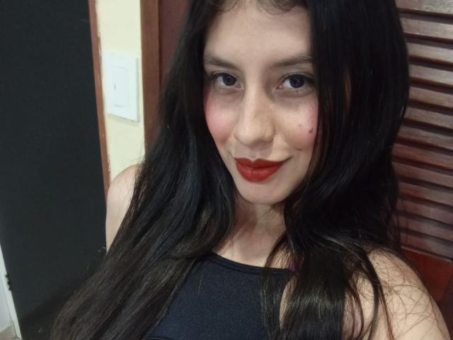 Photograph of MadissonFontaine, a pale-skinned woman with long black hair, red lipstick, and dark eyes, smiling slightly. She's wearing a black sleeveless top, with wooden slats in the background. Fucking sexy.