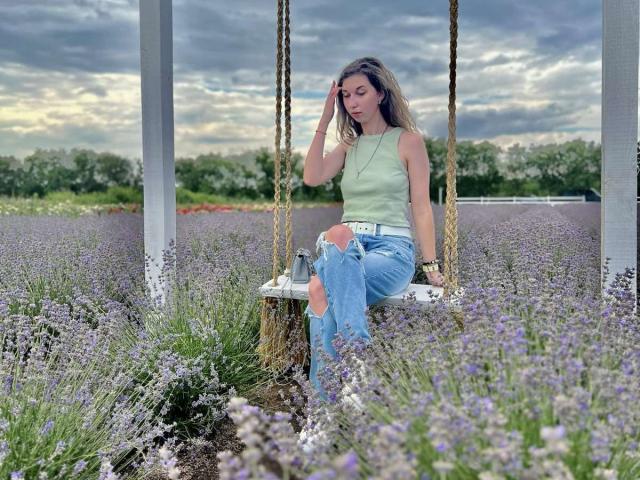 Sanella sits on a wooden swing, wearing a light green tank top and blue jeans, surrounded by a field of purple lavender. She touches her hair, exuding casual allure. Fucking sexy.