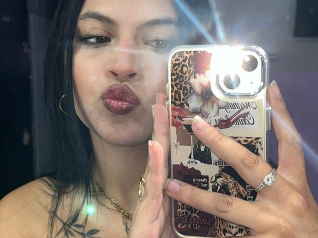 Foto ravvicinata di NyraVanel che si fa un selfie con una custodia per telefono a stampa leopardo. Lei fa la smorfia con le labbra lucide, indossa una collana e un anello d'oro, e tiene il telefono davanti al viso. Un lampo accecante si riflette sullo schermo del telefono..