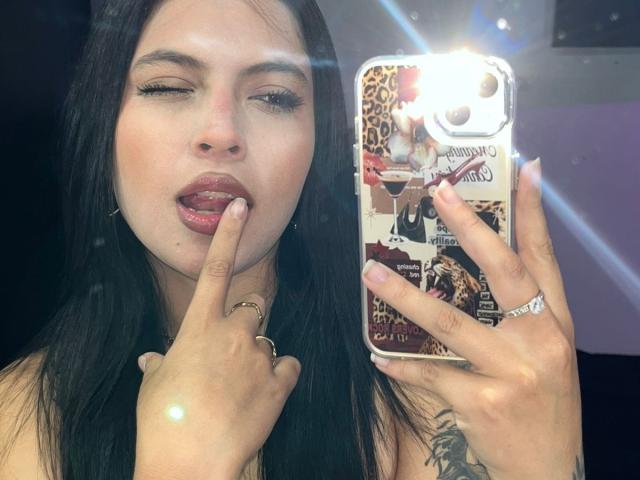 Nahaufnahme von NyraVanel, die ein Telefon mit einer reflektierenden Hülle hält, sich den Finger ableckt und in die Kamera schaut. Halb geschlossene Augen. Heller Blitz, langes, schwarzes Haar, roter Lippenstift.